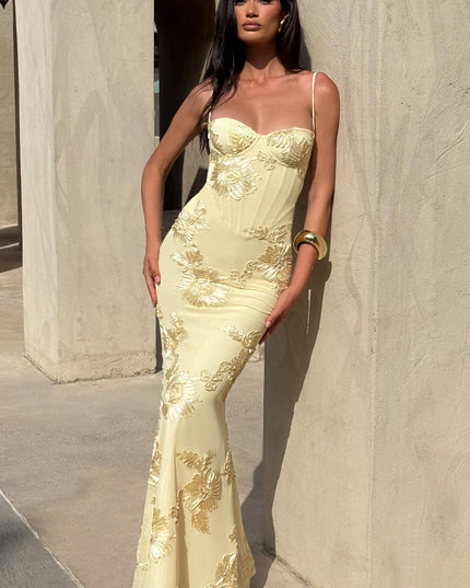 Full-length side view of a woman in a light yellow floral corset maxi dress with gold floral embroidery, posing outdoors.