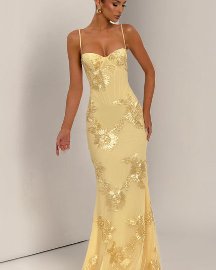 Full-length view of a woman in a light yellow floral corset maxi dress with gold embroidery, posing with one arm outstretched.