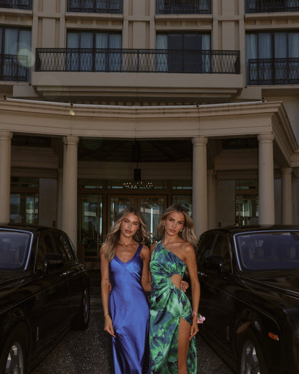 Two women wearing elegant maxi dresses, one in a blue slip style, the other in a green floral one shoulder cut out style, standing between black luxury cars in front of a grand building.
