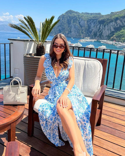 Women's blue and white floral print maxi dress with ruffles and V-neck, worn casually on a sunny balcony overlooking the sea.