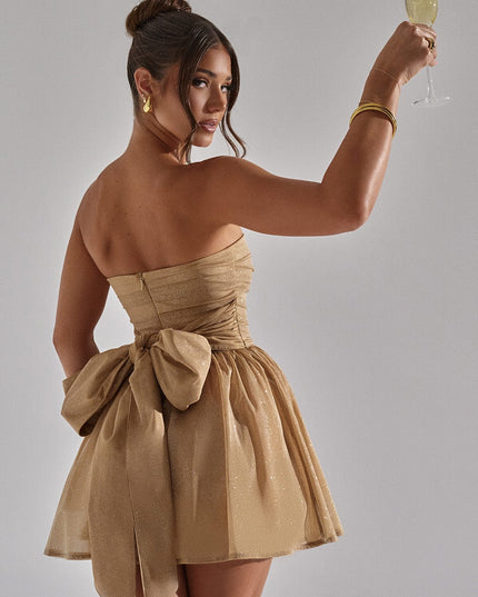 Back view of a woman in a gold strapless party dress, holding a champagne glass, highlighting the bow.