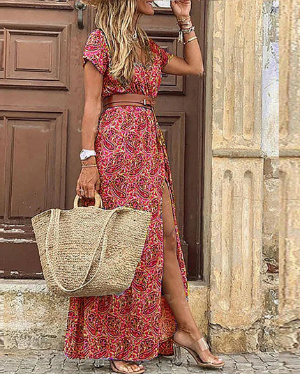 Women's red and pink paisley floral print short sleeve maxi dress with a brown belt and side slit, holding a straw bag.