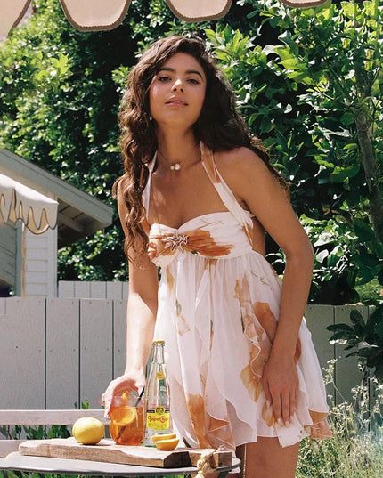 Woman wearing a white summer floral ruffle halter mini dress with orange flowers, enjoying a drink outdoors.