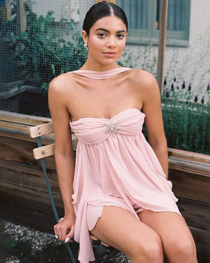 Woman seated wearing a light pink summer ruffle mini dress with a strapless design and silver starfish detail.