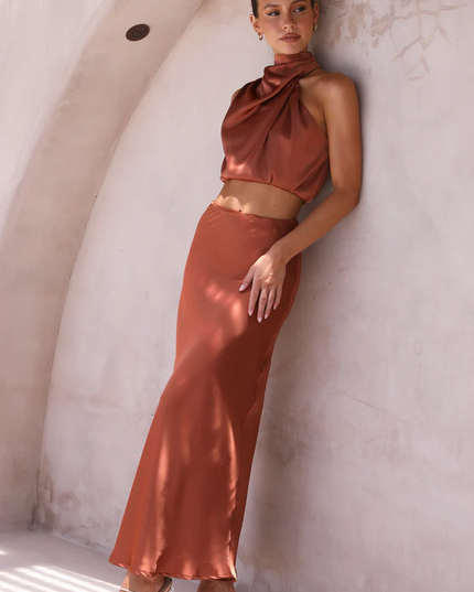 Women's rust orange satin asymmetrical high neck crop top and maxi skirt set, leaning against a wall, showcasing the flowy fabric.