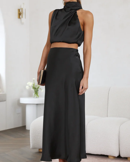 Side view of a woman in a black satin asymmetrical high neck crop top and matching maxi skirt, with black block heels.