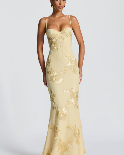 Front view of a woman standing, wearing a light yellow floral lace-up maxi dress with delicate gold flower embroidery and corset detail.