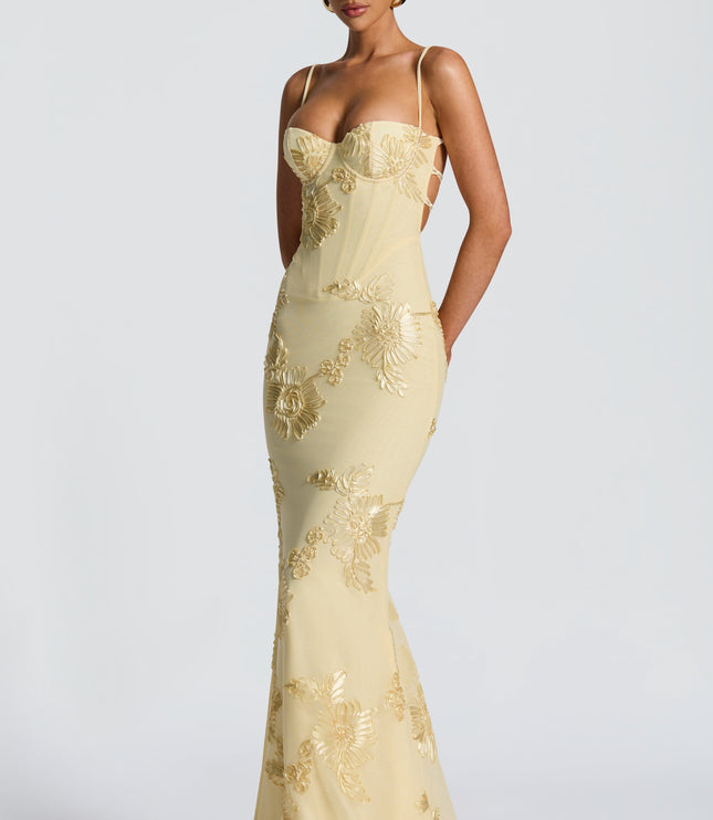 Side view of a woman wearing a light yellow floral lace-up maxi dress, featuring gold embroidered flowers and a silhouette.