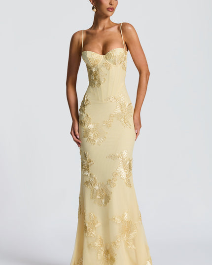 Full front view of a woman standing in a light yellow floral lace-up maxi dress, displaying the gold floral embroidery and bustier top.