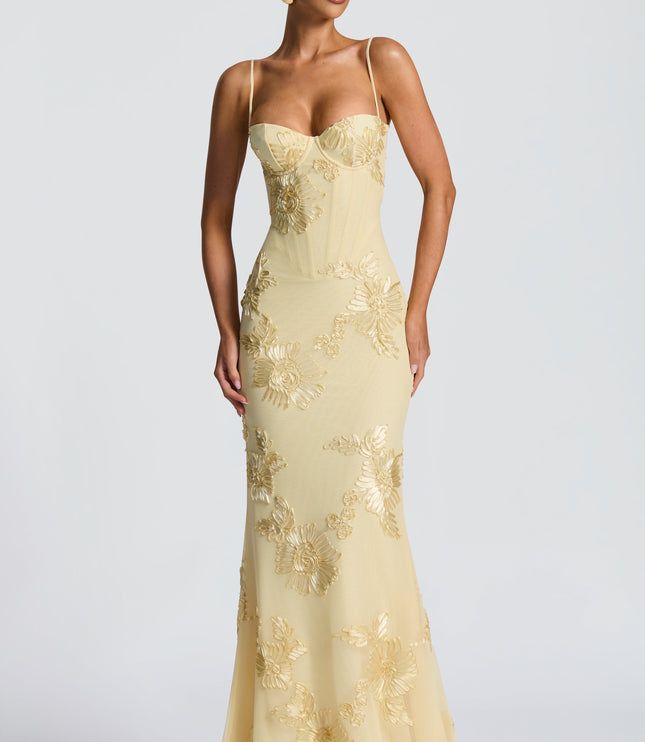 Full front view of a woman standing in a light yellow floral lace-up maxi dress, displaying the gold floral embroidery and bustier top.