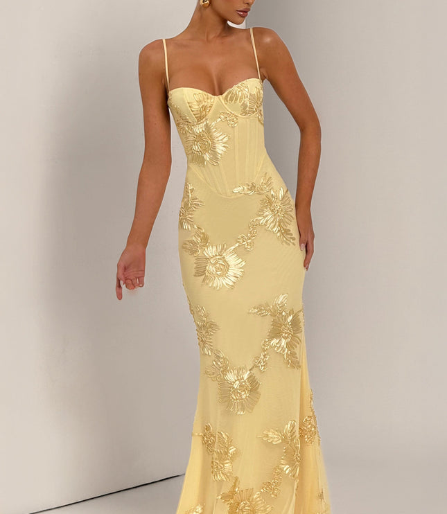 Woman modeling a light yellow floral lace-up maxi dress with gold embroidered flowers, showing the dress's full length and design.