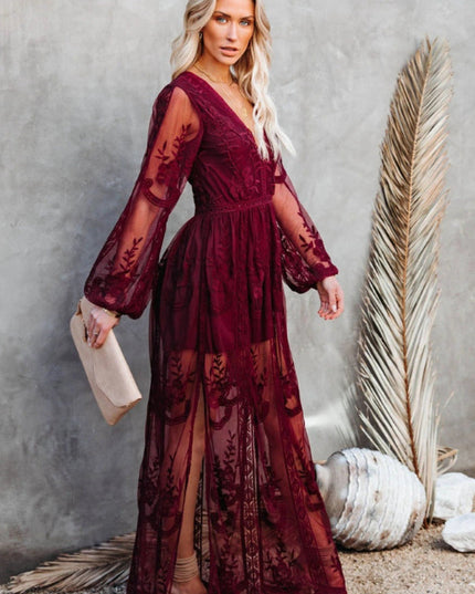 Women's deep burgundy lace long sleeve V-neck maxi dress with sheer skirt and side slit, holding a light clutch.