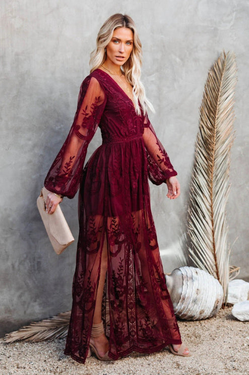Women's deep burgundy lace long sleeve V-neck maxi dress with sheer skirt and side slit, holding a light clutch.