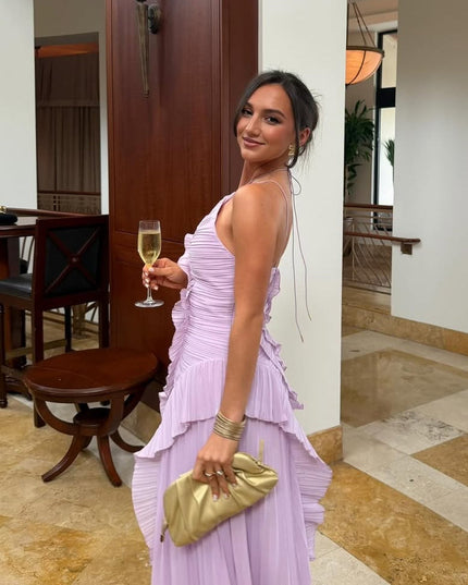 Side profile of the lavender one-shoulder midi dress, showing the pleated skirt and ruffled details, accessorized with a gold clutch.
