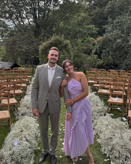 Lavender one-shoulder ruffle midi dress for women, paired with a man in a light green suit, posing outdoors at a wedding.