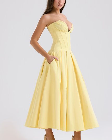 Side profile of a yellow strapless boned bodice pleated midi dress with pockets, paired with nude heels.