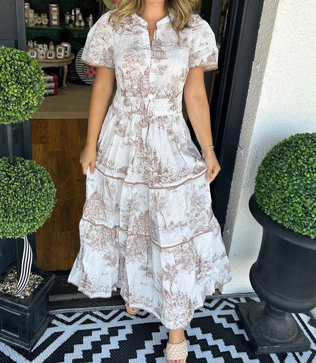 Front view of women's white summer maxi dress with brown scenic print, v-neck, short sleeves, and tiered skirt, with beige slides.