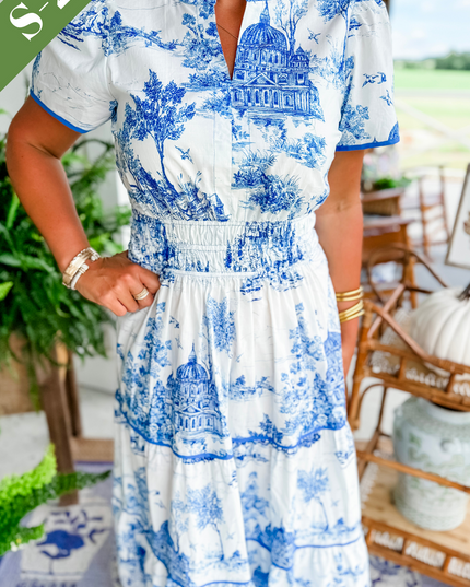 Close-up of women's white summer maxi dress in blue scenic print, showing v-neckline, smocked waist, and tiered skirt detail.