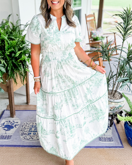 Women's white summer maxi dress with light green scenic print, v-neck, short sleeves, and tiered skirt, posing with hand on skirt.