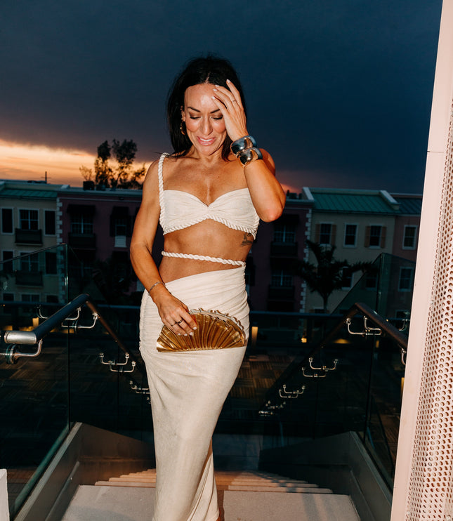 Women's off-white two-piece rope maxi skirt set worn at dusk, featuring the twisted rope details and a gold clutch accessory.