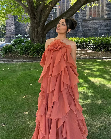 Woman posing outdoors in a coral strapless ruffle layered maxi dress, featuring a prominent floral detail on the bust, against a green lawn and tree.