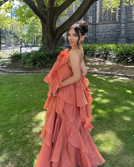 Back view of a woman wearing a coral strapless ruffle layered maxi dress, showcasing the tiered skirt and a subtle back strap detail, in a garden setting.