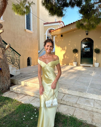 Women's light yellow off-shoulder satin maxi dress with a sweetheart neckline, styled with a white quilted handbag, standing outside a building.