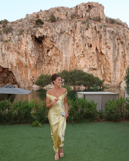 Women's light yellow off-shoulder satin maxi dress with a flowing skirt, worn outdoors with a drink in hand, against a scenic rock and water backdrop.