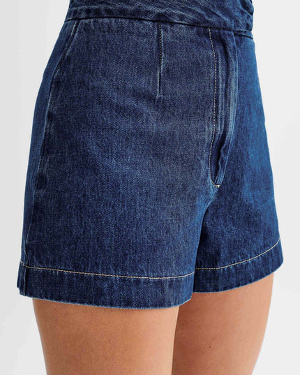 Close-up side view of dark wash denim shorts with a high waist, pleat detail, and contrasting light stitching.