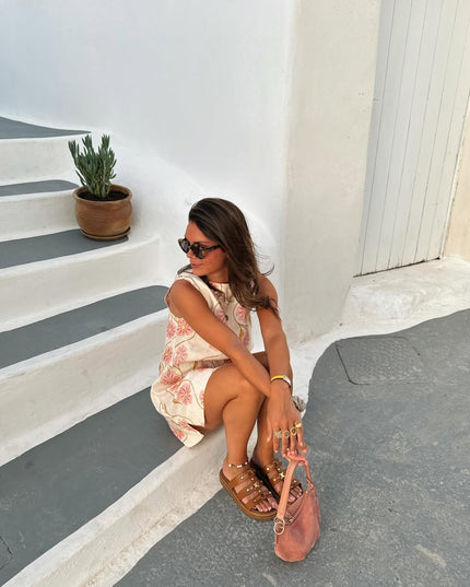 Woman sitting on steps wearing a white summer floral mini dress, brown sandals, and holding a pink bag.