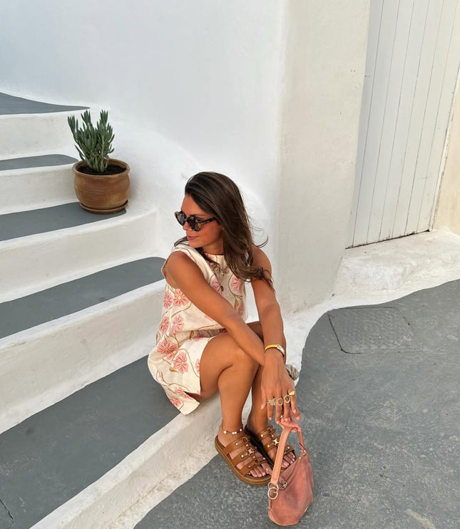Woman sitting on steps wearing a white summer floral mini dress, brown sandals, and holding a pink bag.