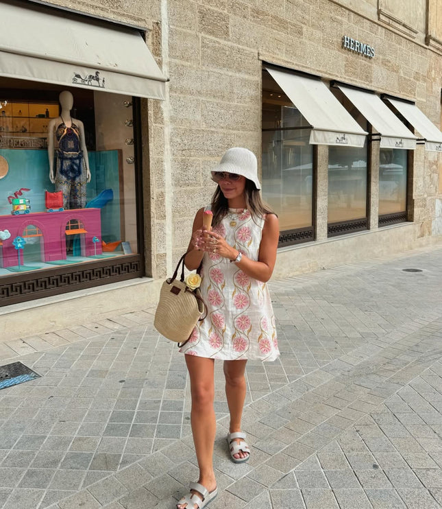 Woman walking on a street, wearing a white summer floral mini dress, white bucket hat, and light grey slides.