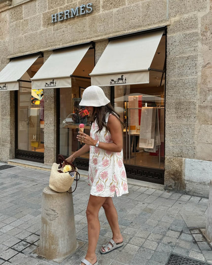 Side view of woman holding ice cream, wearing a white floral sleeveless mini dress and a pleated white bucket hat.