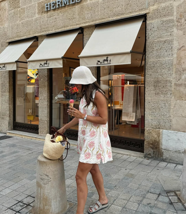 Side view of woman holding ice cream, wearing a white floral sleeveless mini dress and a pleated white bucket hat.