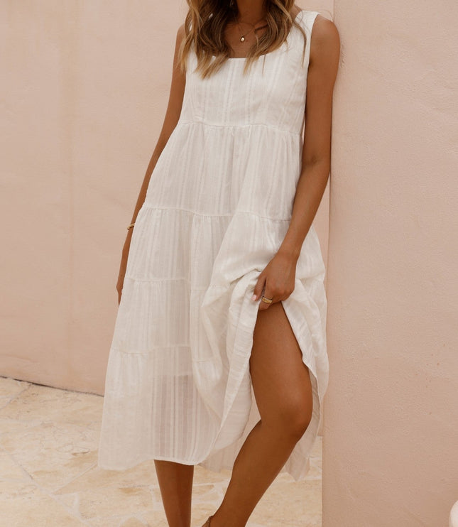 Front view of a woman wearing a white sleeveless tiered midi dress with a square neck and brown flat sandals, leaning against a pink wall.