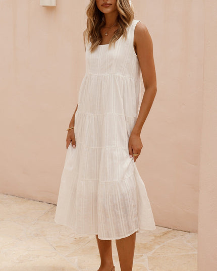 Full body shot of a woman in a white square neck sleeveless tiered midi dress, standing on a light-colored patio.