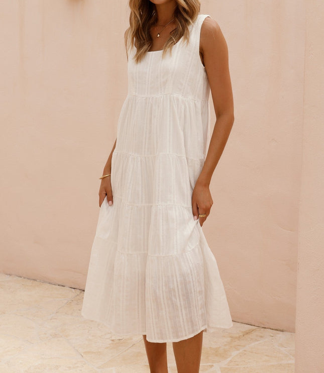 Full body shot of a woman in a white square neck sleeveless tiered midi dress, standing on a light-colored patio.