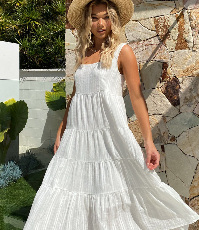 Outdoor shot of the white square neck sleeveless tiered midi dress, with the skirt gently flowing, worn with a straw hat.