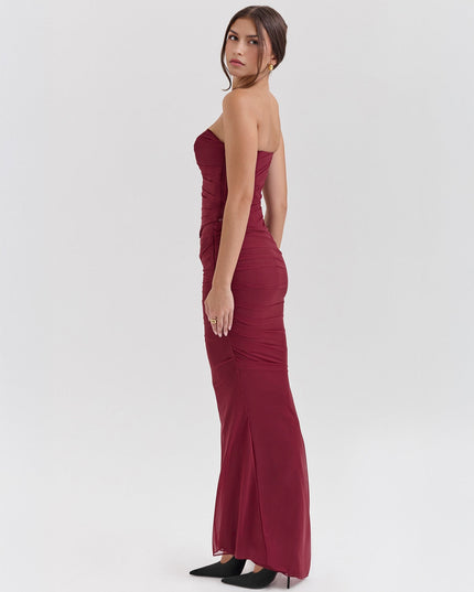 Side profile of a woman in a fitted deep red strapless ruched maxi dress with a sheer overlay, paired with black heels.