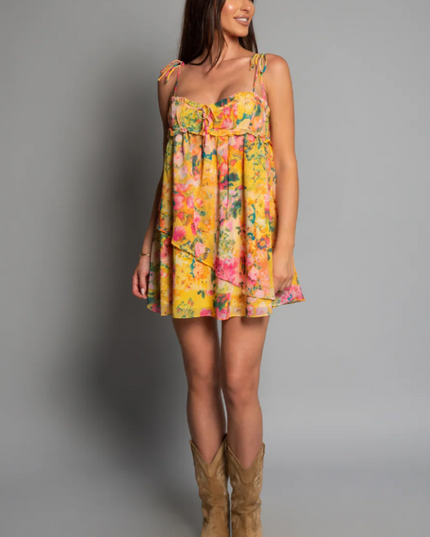 Full length view of a women's yellow floral print mini dress with tie shoulders, paired with cowboy boots.