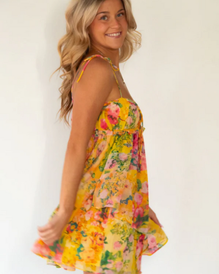 Side view of a blonde woman twirling in a yellow floral print tie shoulder mini dress.