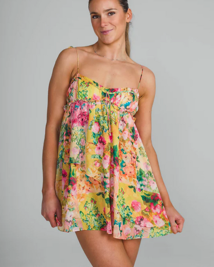Front view of a woman wearing a yellow floral print tie shoulder mini dress, smiling at the camera.