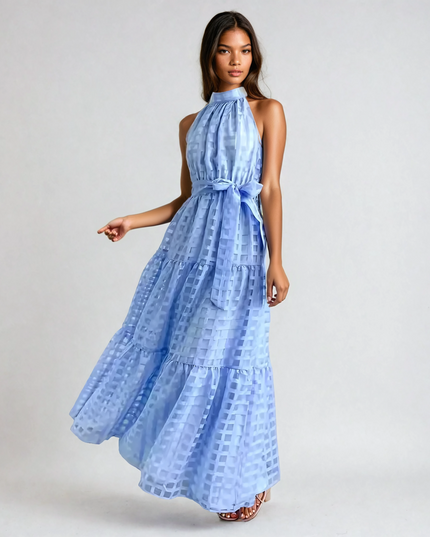 Dark-haired woman wearing a light blue halter neck tiered maxi dress with a subtle checkered pattern and waist tie.