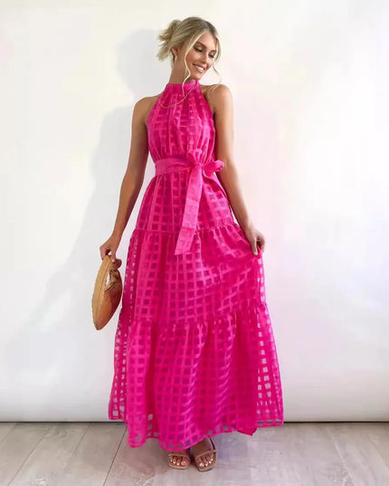 Vibrant pink halter neck tiered maxi dress, showcasing a checkered pattern, waist tie, and a woven bag.